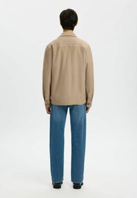 Persoon met kort, donker haar, gekleed in een beige jas, blauwe jeans en zwarte schoenen, staand met de rug naar de camera tegen een witte achtergrond.