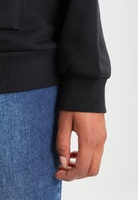 Svart långärmad tröja med ribbade ärmslut, visad med en hand vilande mot sidan. Blå jeans synliga nedanför tröjan.