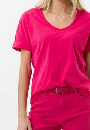 Frau trägt ein leuchtend pinkes kurzärmeliges T-Shirt, das in passende pinke Hosen mit Gürtel gesteckt ist, von den Schultern bis zur Hüfte gezeigt.