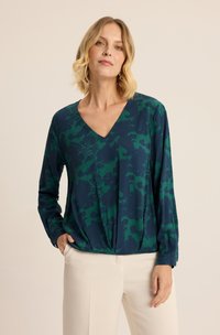 Czarna bluzka z długim rękawem w ciemnozielonym kolorze z kwiatowym wzorem. Posiada głęboki dekolt w serek oraz marszczenie w talii, zestawiona z jasnymi spodniami.