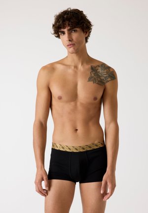 Boxer briefs neri con una vita a motivo dorato. Texture liscia, design aderente e lunghezza fino a metà coscia. Modello che mostra il prodotto.