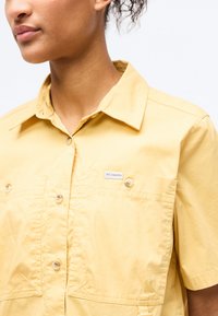 Γυναίκα που φοράει ανοιχτό κίτρινο πουκάμισο με κοντά μανίκια και δύο τσέπες στο στήθος, η μία με μικρή ετικέτα λογότυπου Columbia.