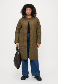 Καπιτονέ μακρύ παλτό σε ελαιόχρωση με στρογγυλή λαιμόκοψη, κουμπιά μπροστά, δύο πλαϊνές τσέπες, φορεμένο πάνω από μαύρη μπλούζα και μπλε φαρδιά τζιν παντελόνια. Μαύρες μπότες και τσάντα tote.