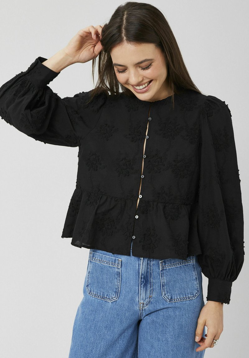 Blouse peplum noire avec motif floral brodé, encolure ronde, fermeture à boutons et manches bouffantes, associée à un jean denim clair.