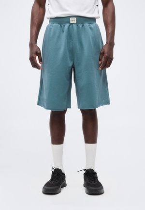 Mies, joka pukeutunut teelisin polvipitkiin shortseihin, valkoisiin sukkiin, mustiin tennareihin ja valkoiseen hihattomaan toppiin, seisoo yksinkertaisen taustan edessä.