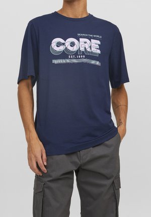 Man wearing navy blue t-shirt with "CORE" and "Search the World EST. 1990" text, paired with gray cargo pants, hand in pocket.