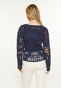 Suéter de crochet azul marino con intrincados patrones florales y de espiral, de mangas largas y diseño corto, combinado con pantalones blancos.
