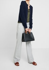 Borsa strutturata nera con manici marroni, abbinata a un cardigan blu navy, un top oliva e pantaloni svasati grigi chiari. Include calzature casual.