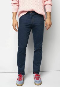 Persoon in donkere blauwe slim-fit jeans, lichtblauwe sneakers met rode veters, en een roze kabelgebreide trui, staand tegen een witte muur.