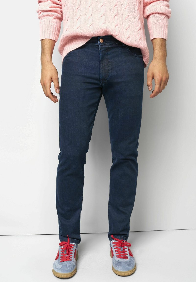 Persoon in donkere blauwe slim-fit jeans, lichtblauwe sneakers met rode veters, en een roze kabelgebreide trui, staand tegen een witte muur.