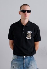 Črna polo majica s kratkimi rokavi, ki na prsih prikazuje risanega medveda, oblečena z modrimi jeans kratkami in temnimi sončnimi očali.