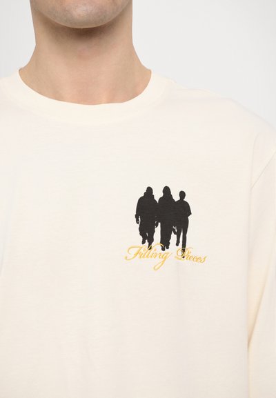T-shirt boje kreme s crnim silueta grafikom tri figure i zlatnim natpisom "Filling Pieces" ispod.