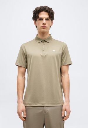 Νέος άνδρας με σκούρα σγουρά μαλλιά, που φοράει μπεζ κοντομάνικη μπλούζα πόλο και παντελόνι που ταιριάζει, στέκεται μπροστά σε απλό λευκό φόντο.