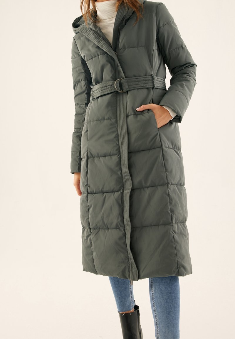 Długa, ciemnozielona puchowa kurtka z paskiem w talii, kapturem i pikowaną fakturą. Posiada boczne kieszenie i prosty krój.