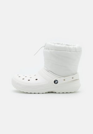 Bota branca com parte superior almofadada, apresentando orifícios de ventilação, sola plana e uma tira lateral. Feita de materiais sintéticos com uma textura suave.