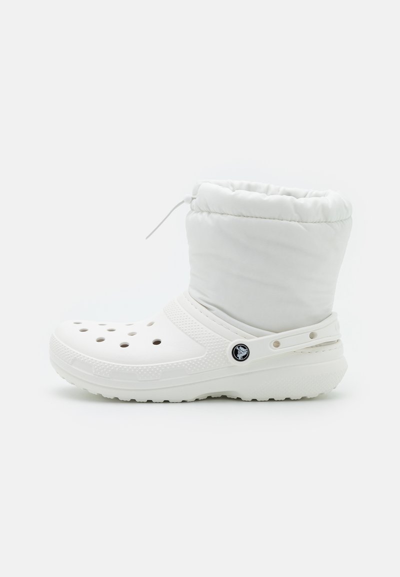 Bota branca com parte superior almofadada, apresentando orifícios de ventilação, sola plana e uma tira lateral. Feita de materiais sintéticos com uma textura suave.