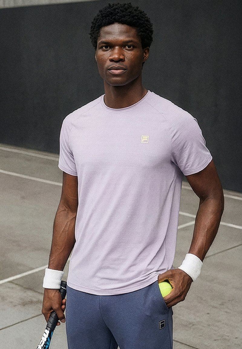 Athlet steht auf dem Tennisplatz und hält einen Schläger und einen Tennisball, trägt ein hellviolettes Shirt und blaue Shorts mit weißen Handgelenkbandagen.