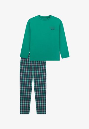 Ragazzo sorridente a piedi nudi in piedi, che indossa una camicia verde a maniche lunghe e pantaloni a quadri verdi e blu su uno sfondo bianco semplice.