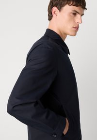 Casaco azul-marinho com um design ajustado, colarinho clássico, detalhes em botão e uma textura suave, exibindo uma vista de perfil lateral.