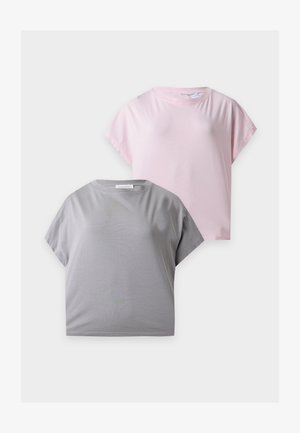 Vrouw met lang bruin haar, die een los grijs topje draagt, wijd uitlopende blauwe spijkerbroek en witte sneakers, staand met één hand in haar zak tegen een witte muur.