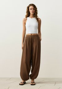 Femme aux cheveux bouclés portant un crop top blanc sans manches, un pantalon marron ample et des sandales noires, debout devant un fond clair uni.