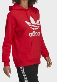 Röd Adidas hoodie med en stor vit logotyp och trebladsmönster. Har en framficka och dragsko i huvan. Tillverkad av bomull.
