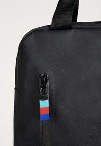 Sac en tissu noir avec surface texturée, fermeture éclair latérale munie d'un tirette multicolore, et poignées supérieures robustes pour le transport.