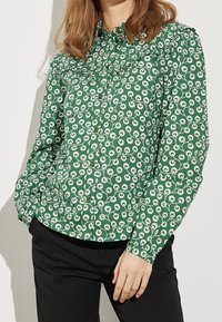 Chemise verte à boutons avec un motif floral blanc, dotée d'un col et de manches longues, fabriquée dans un tissu doux.