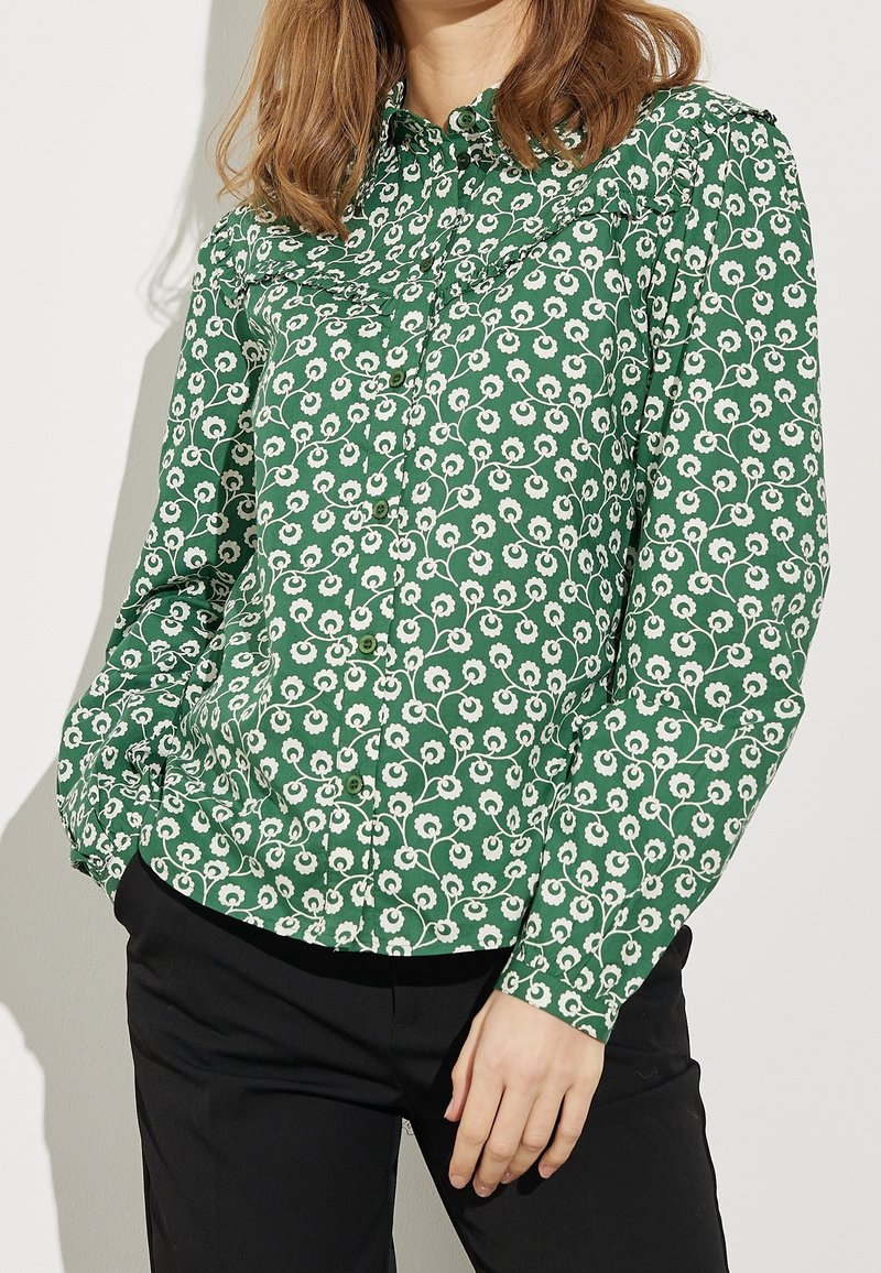 Chemise verte à boutons avec un motif floral blanc, dotée d'un col et de manches longues, fabriquée dans un tissu doux.