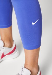Legginsy Nike o długości 7/8 w żywym królewskim niebieskim kolorze, z białym logo po boku. Materiał jest gładki i elastyczny. Szczegółowo wykończony dół.