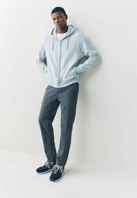 Világosszürke cipzáras kapucnis pulóver húzózsinórokkal, sötétszürke nadrággal és fekete sportcipőkkel, szürke részletekkel. Végig sima textúrák.