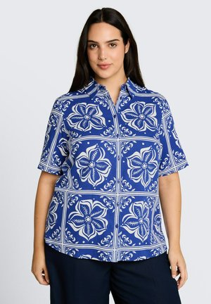 Frau mit langen dunklen Haaren, die ein blaues kurzärmeliges Hemd mit weißen großen Blumen- und geometrischen Mustern trägt.