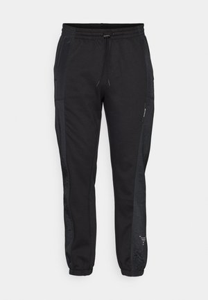 Schwarze Jogginghose mit elastischem Bund, Kordelzug und seitlichen Taschen. Mit strukturierten schwarzen Seitenpaneelen und elastischen Bündchen an den Knöcheln.