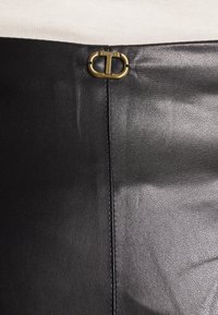 Black leather skirt with central seam and small gold oval buckle engraved with "TWINSET" at the waistband against a beige top background.