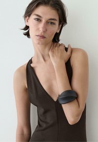 Frau mit kurzen braunen Haaren, die ein ärmelloses dunkelbraunes Oberteil und ein klobiges schwarzes Armband am rechten Arm trägt, berührt ihre linke Schulter.