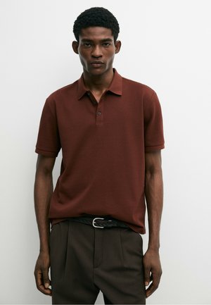 Jonge man met kort krullend haar, die een bruin poloshirt met korte mouwen en donkere broek draagt tegen een effen witte achtergrond.