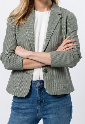Femme aux bras croisés portant un blazer gris texturé, une blouse blanche et un jean bleu sur un fond uni.