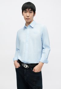 Jasnoniebieska koszula w paski na guziki z ostro zakończonym kołnierzykiem, noszona z ciemnymi jeansami z haftowanymi naszywkami i srebrną sprzączką paska.