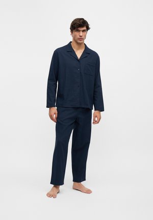 Joven de pie descalzo, vestido con una blusa de manga larga azul oscuro abotonada y pantalones anchos a juego sobre un fondo blanco.