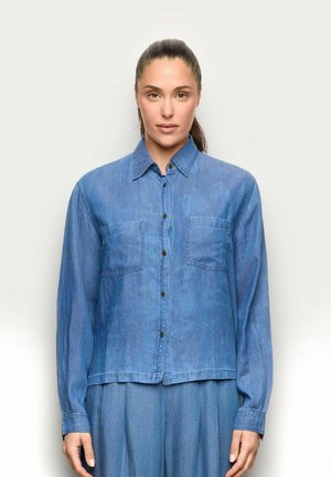 Femme aux cheveux foncés attachés en queue de cheval, portant une chemise en denim bleu clair boutonnée et un pantalon assorti, face à l'avant sur un fond uni.
