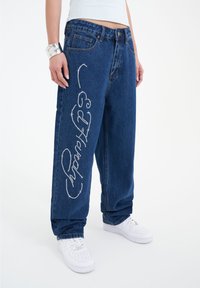 Top corto azul claro, jeans de mezclilla oscuros de corte holgado con detalles de strass en la pierna izquierda, combinados con zapatillas blancas.