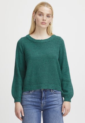 Pull en laine vert avec un col rond, motif texturé et manches côtelées. Associé à un jean en denim bleu, mettant en valeur une coupe décontractée.