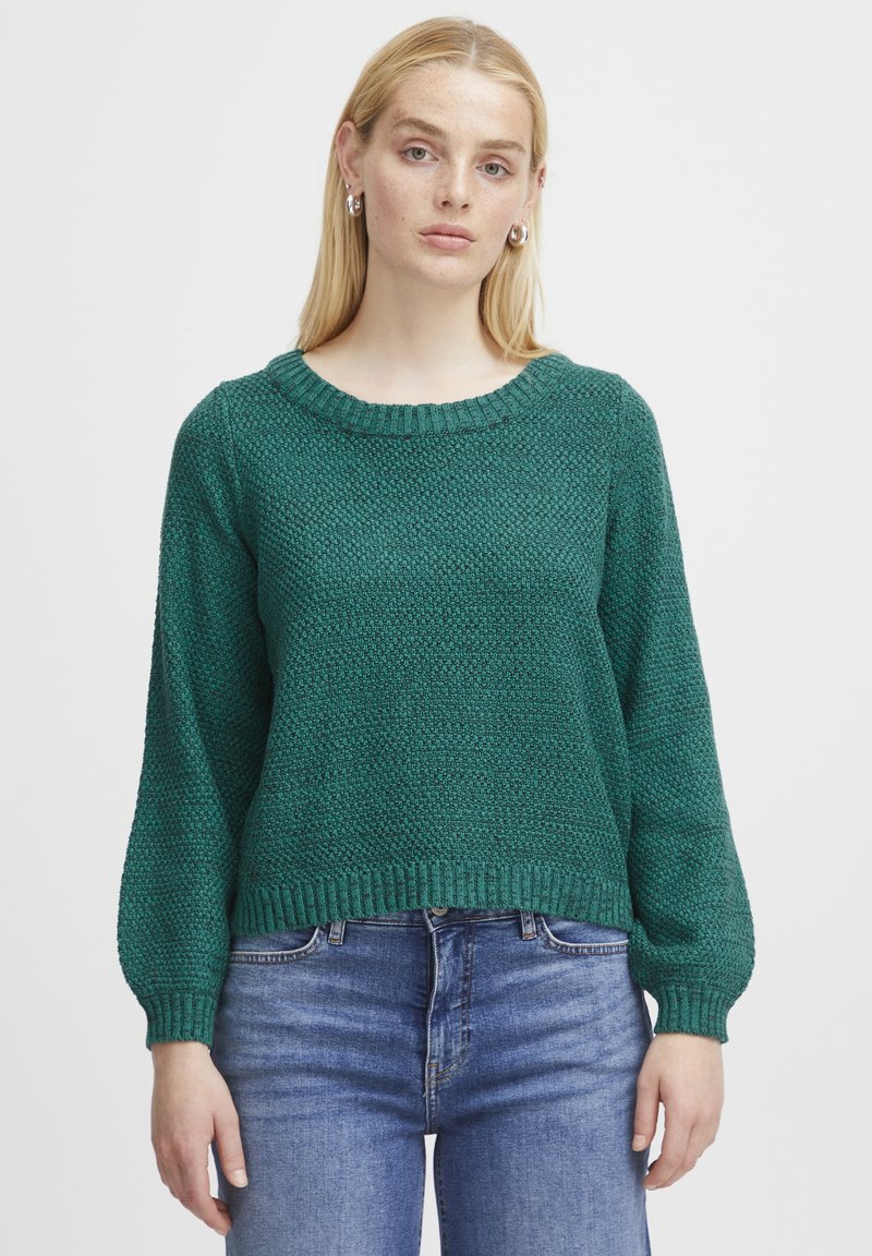 Grüner gestrickter Pullover mit rundem Ausschnitt, strukturiertem Muster und gerippten Bündchen. Kombiniert mit blauen Jeans aus Denim, die eine lockere Passform zeigen.
