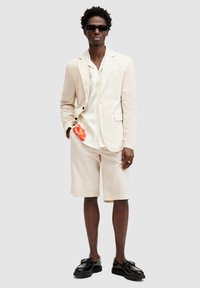 Giacca di costume beige chiaro sopra una camicia bianca, abbinata a pantaloni corti coordinati. Dettaglio aggiuntivo: un fiore arancione spunta dalla tasca. Occhiali da sole neri e scarpe.