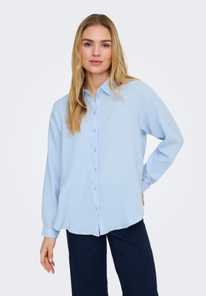 Vaaleatukkainen nainen, joka käyttää vaaleansinistä pitkähihainen nappipaitaa ja tummia housuja, seisoo yhdellä kädellä lanteilla yksinkertaista taustaa vasten.