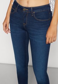 Vaqueros de mezclilla azul oscuro con corte ajustado, diseño de cinco bolsillos, costuras de cobre y un botón metálico en la cintura.