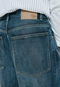 Traperice s tamnim plavim pranjem, sa dva stražnja džepa i smeštnom kožnom etiketom na struku s utisnutim logotipom.