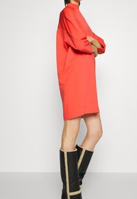 Koralfarbenes oversized Kleid mit langen Ärmeln und hohem Kragen, aus weichem Stoff gefertigt. Getragen mit knielangen schwarzen Stiefeln mit cremefarbenen Akzenten.