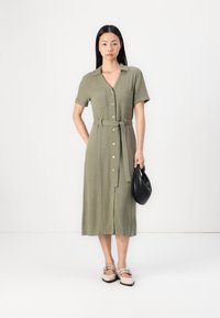 Robe olive verte à manches courtes, avec des boutons, ceinturée à la taille, deux poches poitrine et une longueur midi, accompagnée d'un sac à main noir.
