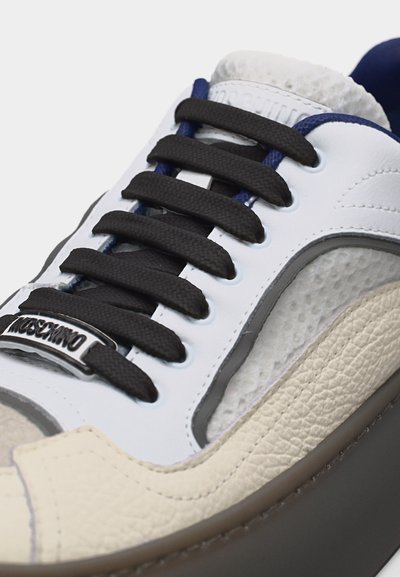Λευκό και μπεζ αθλητικό παπούτσι με μαύρα κορδόνια και μεταλλικό λογότυπο Moschino στο μπροστινό μέρος, που δείχνει τμήματα από δέρμα με υφή και λείο δέρμα.